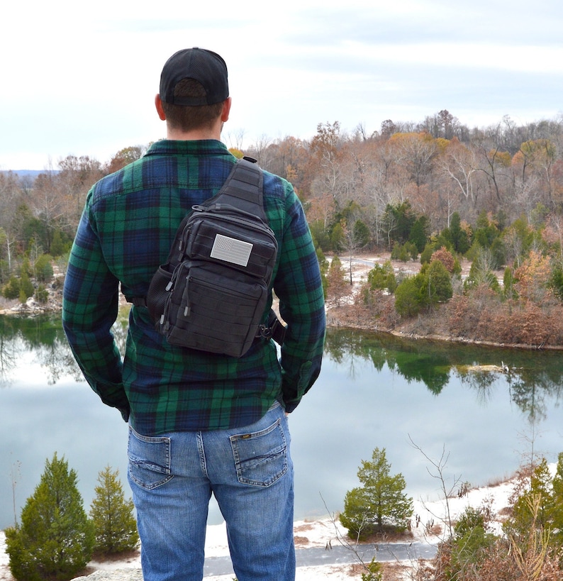 May include: A person wearing a black and green plaid shirt and blue jeans is standing with their back to the camera. They are wearing a black tactical sling bag with a gray patch on the front. The person is looking out at a lake in the distance.