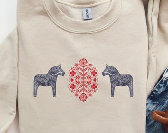 May include: A beige crewneck sweatshirt featuring a graphic print of two dark gray Dala horses facing a central red floral pattern. The design is centered on the chest of the garment, which has long sleeves and a ribbed collar, cuffs, and hem.