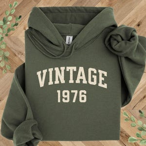 May include: An olive green hooded sweatshirt with the word "VINTAGE" and the year "1976" printed in a cream-colored, collegiate-style font. The hoodie has a drawstring and a rolled-up sleeve.