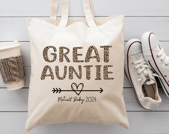 May include: A cream-colored tote bag with the words "GREAT AUNTIE" in leopard print. Below the text is an arrow with a heart and the text "Metcalf Baby 2024". White sneakers and a coffee cup are next to the bag.