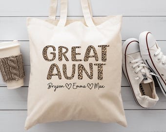 May include: Cream-colored tote bag with the words "GREAT AUNT" in leopard print. Below the text are the names "Bryson", "Emma", and "Max" with heart symbols. A white coffee cup and white sneakers are next to the bag.
