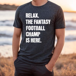 Op de afbeelding: Een donkergrijs T-shirt met de tekst "Relax. The Fantasy Football Champ Is Here." in wit gedrukt.