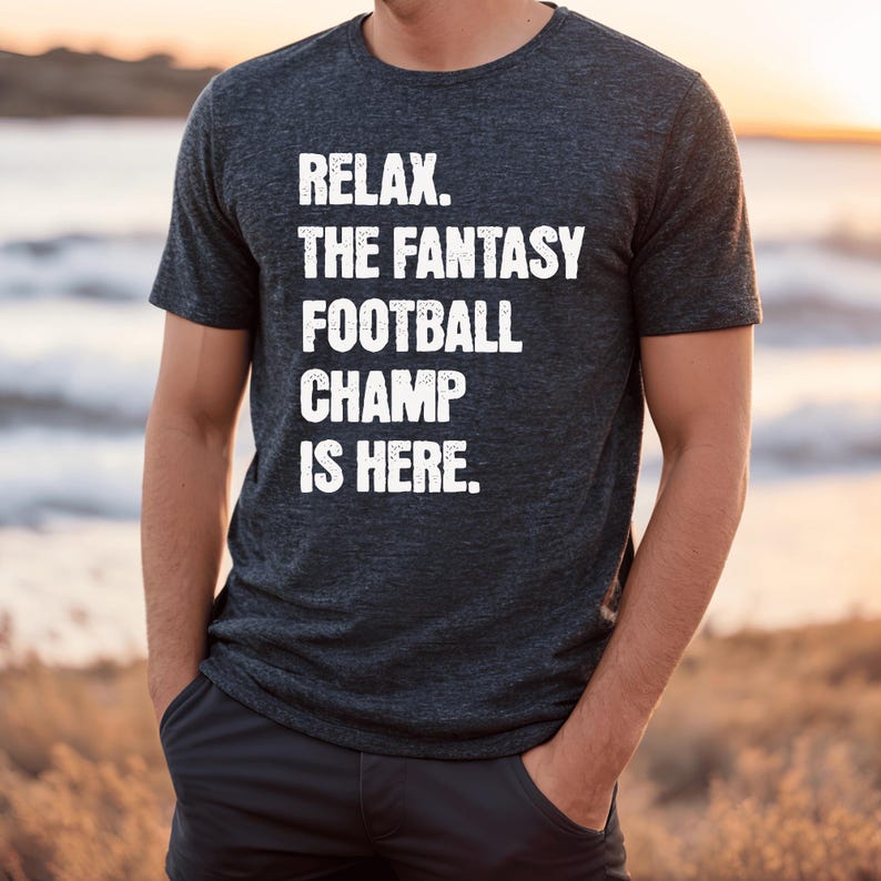 Op de afbeelding: Een donkergrijs T-shirt met de tekst "Relax. The Fantasy Football Champ Is Here." in wit gedrukt.
