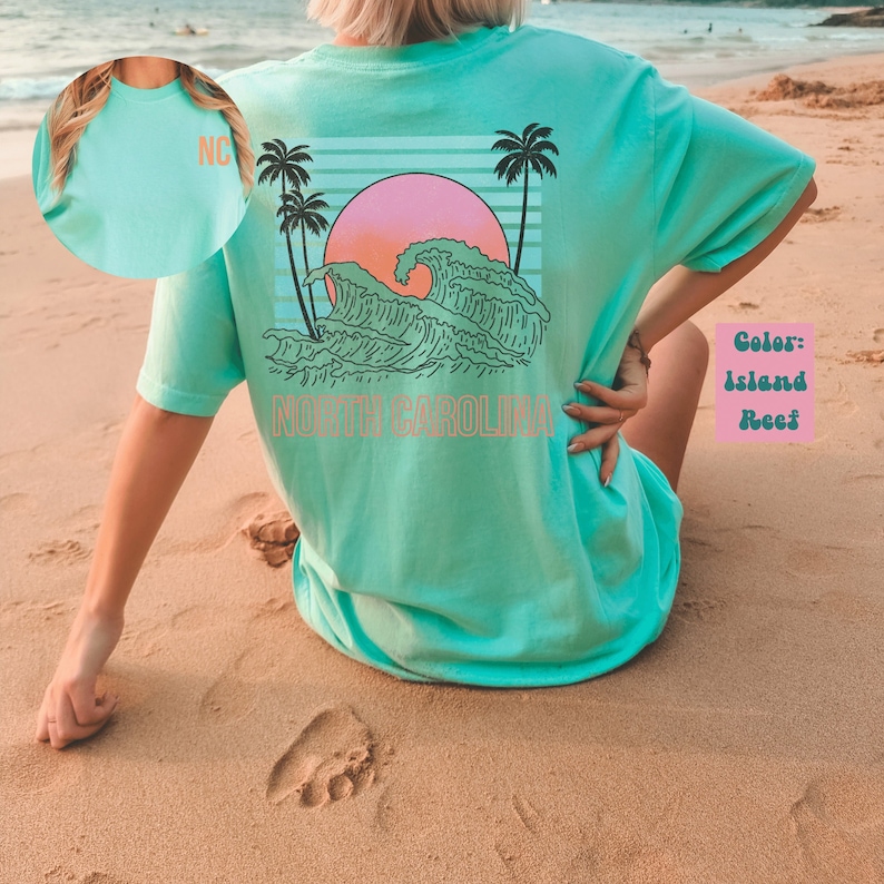 May include: A turquoise blue t-shirt with a graphic of a sunset over a wave and the text "NORTH CAROLINA". The graphic is in shades of pink, orange, and green. The t-shirt is being worn by a person sitting on a sandy beach.