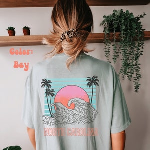 May include: A light blue t-shirt with a graphic of a wave crashing against a sunset with palm trees on either side. The text "NORTH CAROLINA" is printed below the graphic.