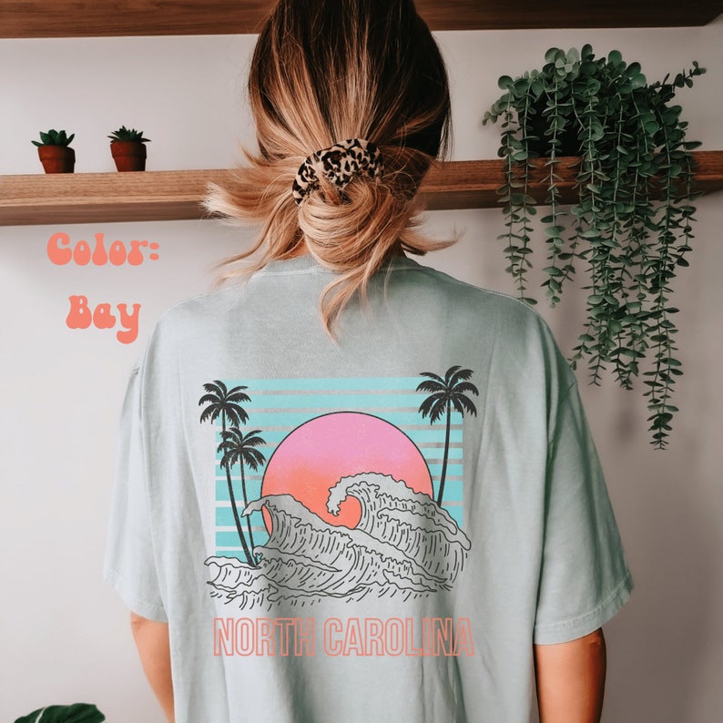 May include: A light blue t-shirt with a graphic of a wave crashing against a sunset with palm trees on either side. The text "NORTH CAROLINA" is printed below the graphic.