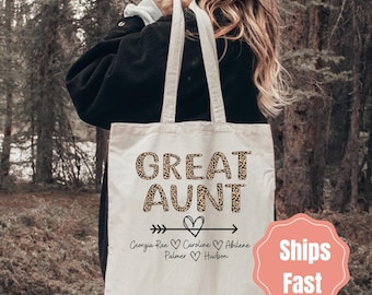 May include: Cream-colored tote bag with the words "GREAT AUNT" in a leopard print font. Below the text is an arrow with a heart, and names. The bag is held by a person in a black jacket.
