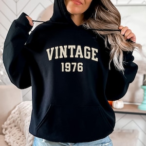 May include: A black hooded sweatshirt featuring the word VINTAGE in a distressed, off-white, arched font above the year 1976 in a matching distressed, off-white, block font. The hoodie includes a front kangaroo pocket and black drawstrings.