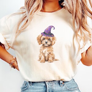 May include: A light beige sweatshirt featuring a watercolor illustration of a small, brown dog wearing a purple witch hat with gold stars. The dog is sitting and looking forward. The sweatshirt is a neutral color.