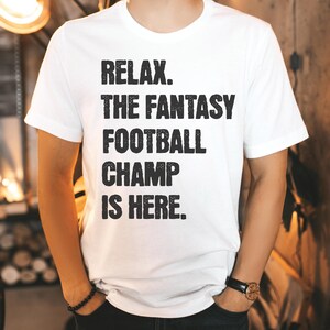 Op de afbeelding: Wit T-shirt met zwarte tekst die "Relax. The Fantasy Football Champ Is Here." luidt.