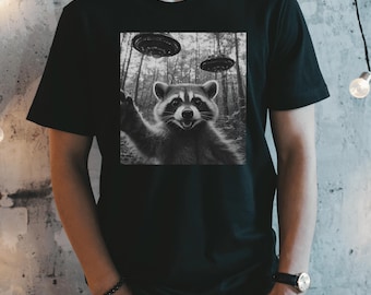 Pode incluir: Uma camiseta preta com um gráfico de um guaxinim acenando para três discos voadores em uma floresta em preto e branco.