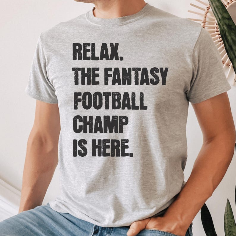 Op de afbeelding: Een grijs T-shirt met de tekst "Relax. The Fantasy Football Champ Is Here." in zwart gedrukt.