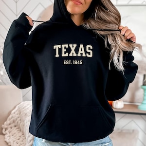 May include: Black hooded sweatshirt with the word "TEXAS" in white block letters above "EST. 1845". The sweatshirt has a front pocket and a drawstring hood. The person is wearing blue jeans.