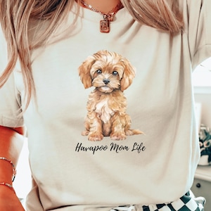 May include: Cream-colored t-shirt featuring a watercolor illustration of a Havapoo puppy. The puppy has light brown fur and is sitting. The text "Havapoo Mom Life" is printed in black script below the puppy.