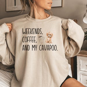 May include: A beige sweatshirt with the text "Weekends, Coffee, and My Cavapoo." A cartoon image of a dog and a coffee cup are also on the sweatshirt.