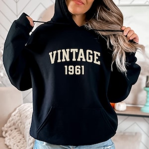 May include: Black hooded sweatshirt featuring the word "VINTAGE" above the year "1961" in a cream-coloured, block font. The sweatshirt has a front pocket and a drawstring hood.