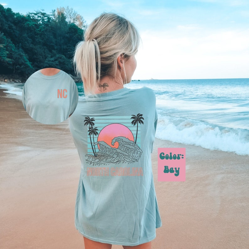 May include: A light blue long-sleeved t-shirt with a graphic of a sunset over waves and palm trees. The text "NORTH CAROLINA" is printed below the graphic. The shirt has a light pink and blue color scheme. The color of the shirt is "Bay".