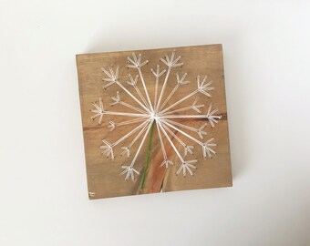 May include: Wooden square art piece featuring a dandelion design. White string is strung between nails to create the flower's head, with a green stem. The wood has a natural, light brown finish.