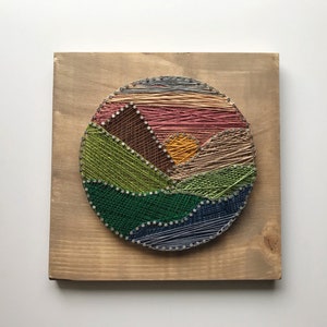 May include: A colorful string art piece depicting a landscape scene. The artwork features a circular design with various colored threads, including green, blue, pink, and yellow, creating a mountain and sunset effect. The art is mounted on a wooden board.