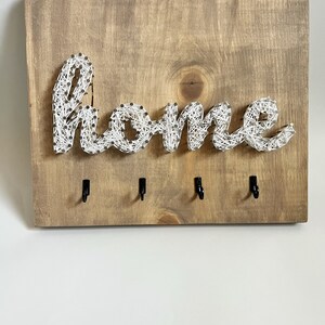 Puede incluir: Portallaves de madera con la palabra "home" hecha con hilo blanco y clavos. El tablero de madera marrón tiene cuatro ganchos negros. Un elemento decorativo y práctico para la organización del hogar.
