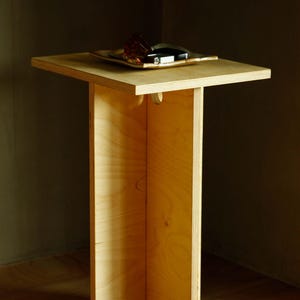 May include: A light-colored, wooden side table with a square top and a single, central support. The table has a natural wood grain finish. A small tray with items sits on top. The table is in a corner of a room.