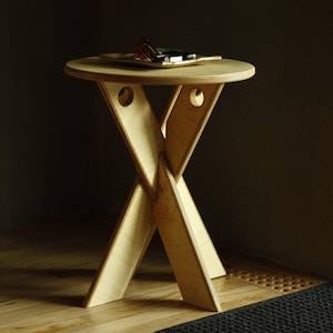 May include: A round light-colored plywood side table featuring a cross-legged base design with circular cutouts near the top of each leg. The table has a smooth, flat circular top surface with a small, dark, rectangular object resting on it.
