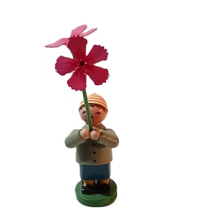 Könnte beinhalten: Eine Holzfigur einer Person, die eine rosa Blume hält. Die Figur trägt eine grüne Jacke, eine blaue Hose und eine gelb-weiß gestreifte Mütze.