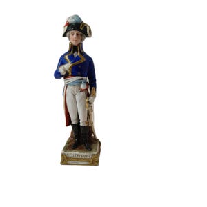 Könnte beinhalten: Porzellanfigur eines Militäroffiziers in einer blauen Jacke mit goldenen Verzierungen, weißer Hose und Dreispitz. Die Figur steht auf einem rechteckigen Sockel mit dem Namen "KELLERMANN". Der Offizier hält ein Schwert.