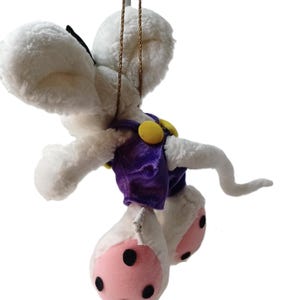 Könnte beinhalten: Ein weißes Plüschmaus-Ornament, das an einer goldenen Kette hängt. Die Maus trägt ein lila Outfit mit gelben Knöpfen und hat rosa Füße mit schwarzen Punkten. Die Maus hat große, flauschige Ohren.