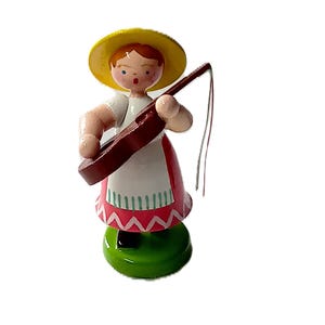 Könnte beinhalten: Holzfigur einer Person, die ein Saiteninstrument spielt. Die Figur trägt einen gelben Hut, ein weißes Oberteil und einen roten Rock mit einem weißen und rosa Zickzackmuster. Der Sockel ist grün.