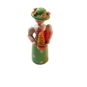 Könnte beinhalten: Eine Holzfigur einer Frau, die einen grünen Hut mit gelben Blumen und ein grünes Kleid mit weißen Punkten trägt. Sie hält einen Topf mit Blumen mit rosa, gelben und roten Blüten.