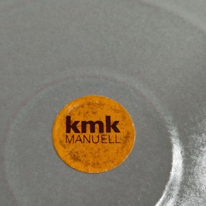 Op de afbeelding: Een close-up van een ronde, oranje sticker met de tekst "kmk MANUELL" in bruine letters.