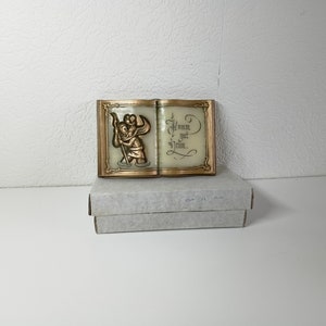 Op de afbeelding: Een goudkleurige, open boekvormige wanddecoratie met een reliëfbeeld van twee engelen. Het boek is gesloten en heeft de woorden "Komm get heim." op de rechterkant.