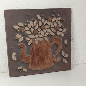 Op de afbeelding: Een bruine keramische tegel met een mozaïekontwerp van een bruine theepot met witte bloemen. De theepot is gevuld met witte bloemen en bladeren, de achtergrond is donkerbruin.