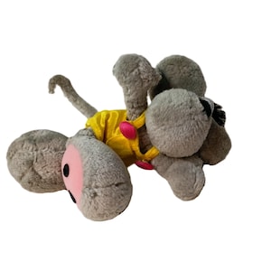 Könnte beinhalten: Ein graues Plüschmaus-Spielzeug mit großen Ohren und einem langen Schwanz. Die Maus trägt ein gelbes Hemd mit rosa Knöpfen. Die Ohren der Maus haben rosa Innendetails und schwarze Augen. Das Spielzeug ist für Kinder konzipiert.