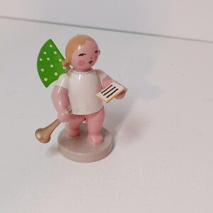 Könnte beinhalten: Holzfigur eines Engels mit einem grünen Flügel mit weißen Punkten, der eine goldene Trompete und ein kleines Blatt Papier hält. Der Engel hat rosa Haut, ein weißes Hemd und steht auf einem runden Sockel.
