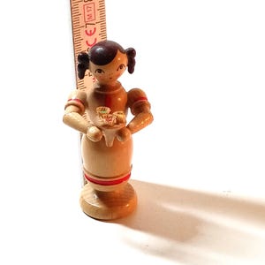 Könnte beinhalten: Eine Holzfigur eines Mädchens mit braunen Haaren und einem roten Kleid, das einen Teller mit Gebäck hält. Die Figur ist etwa 18 cm groß und aus Holz gefertigt.