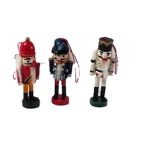 May include: Three miniature wooden nutcracker figurines. The nutcrackers are painted in red, white, blue, and black, with gold accents. Each figure has a different uniform and is standing on a round base. They are Christmas ornaments.