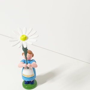 Könnte beinhalten: Holzfigur einer Person, die eine große weiße Gänseblume mit gelber Mitte hält. Die Figur trägt ein blaues Kleid, eine weiße Schürze und steht auf einem grünen Sockel. Die Figur hat braune Haare und rosa Arme.