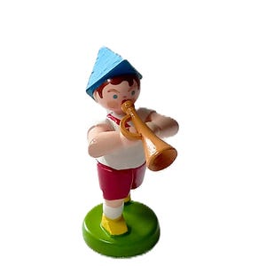 Könnte beinhalten: Holzfigur eines Jungen, der Trompete spielt. Der Junge trägt einen blauen Hut, ein weißes Hemd, rote Shorts und gelbe Schuhe. Die Figur ist auf einem grünen, kreisförmigen Sockel montiert. Die Trompete ist hellbraun.