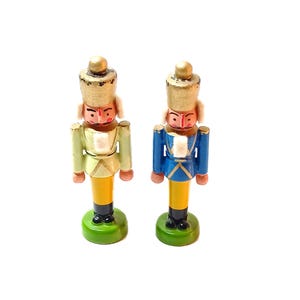 May include: Two miniature wooden nutcracker figurines. One is painted in a light green uniform, the other in blue. Both have gold-coloured hats, red faces, and yellow legs. They stand on green circular bases.