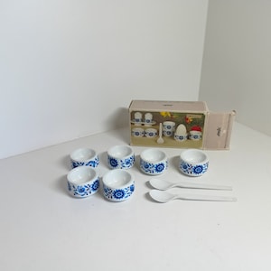Peut inclure: Ensemble de six coquetiers en céramique blanche avec des motifs floraux bleus et deux cuillères en plastique blanc. Les coquetiers sont dans une boîte avec le mot "Simply" dessus.