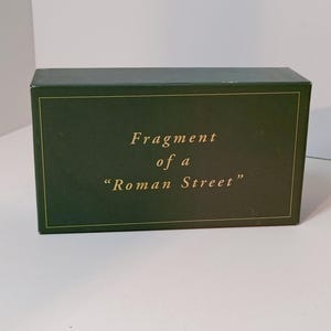 Könnte beinhalten: Eine dunkelgrüne rechteckige Schachtel mit goldfarbenen Buchstaben. Der Text auf der Schachtel lautet "Fragment of a 'Roman Street'". Die Schachtel steht auf einer weißen Oberfläche.
