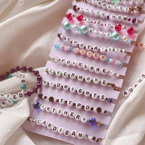 May include: A collection of 14 beaded bracelets with different album titles from Taylor Swift's discography. The bracelets are arranged in rows on a white background. The album titles include Taylor Swift, Fearless, Speak Now, 1989, Reputation, Lover, Folklore, Evermore, Midnights, 1989, Anti-Hero, Cardigan, August, and Willow.