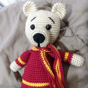 May include: A crocheted teddy bear wearing a red and yellow striped scarf. The bear has black eyes and a black nose.