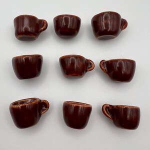 May include: Nine small, brown ceramic teacups with handles are arranged on a white surface. The cups have a glossy finish and a classic design. The teacups are a rich, dark brown color.