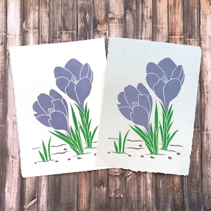 Op de afbeelding: Twee prints van een gestileerde illustratie van paarse krokussen met groene bladeren op een witte achtergrond. De prints zijn op verschillend gekleurd papier, een wit en een lichtblauw.
