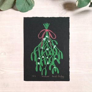 Op de afbeelding: Een zwart-witte illustratie van een tak mistel met groene bladeren en witte bessen. De mistel is vastgebonden met een rood lint en de tekst "mistletoe" staat onder de afbeelding.