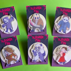 May include: A collection of six acrylic "Slasher Pin" badges, each featuring a different horror movie character. The pins are mounted on purple and black backing cards with the text "Slasher Pin". The characters are in various colors, including purple, brown, and blue.