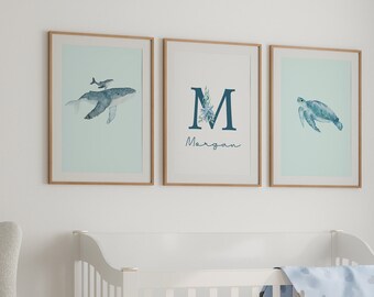 May include: A white crib with a blue and white patterned blanket. Three framed prints hang on the wall above the crib. The prints feature a whale, the letter M with the name "Morgan" written below, and a sea turtle.